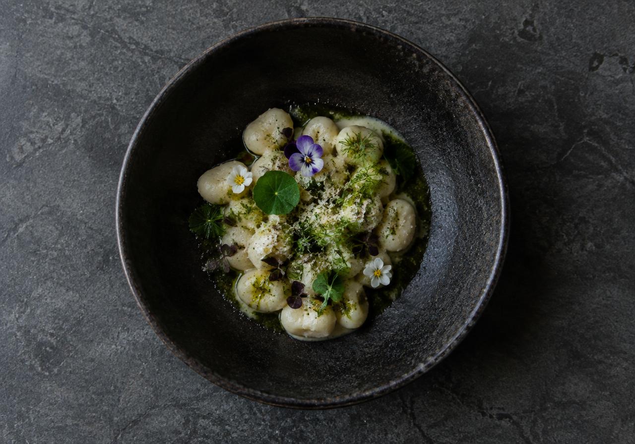 Plated gnocchi with herbs and edible flowers in a dark ceramic bowl, from the Juno Kookt website